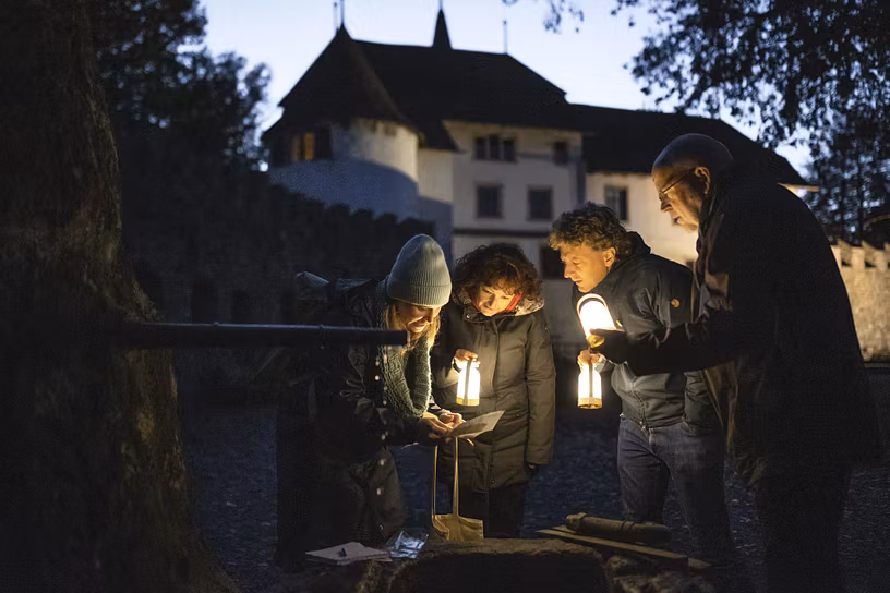 Vier Personen stehen nachts mit Lampfen vor einem Brunnen und studieren einen Plan. Im Hintergrund Schloss Hallwyl in der Dämmerung.