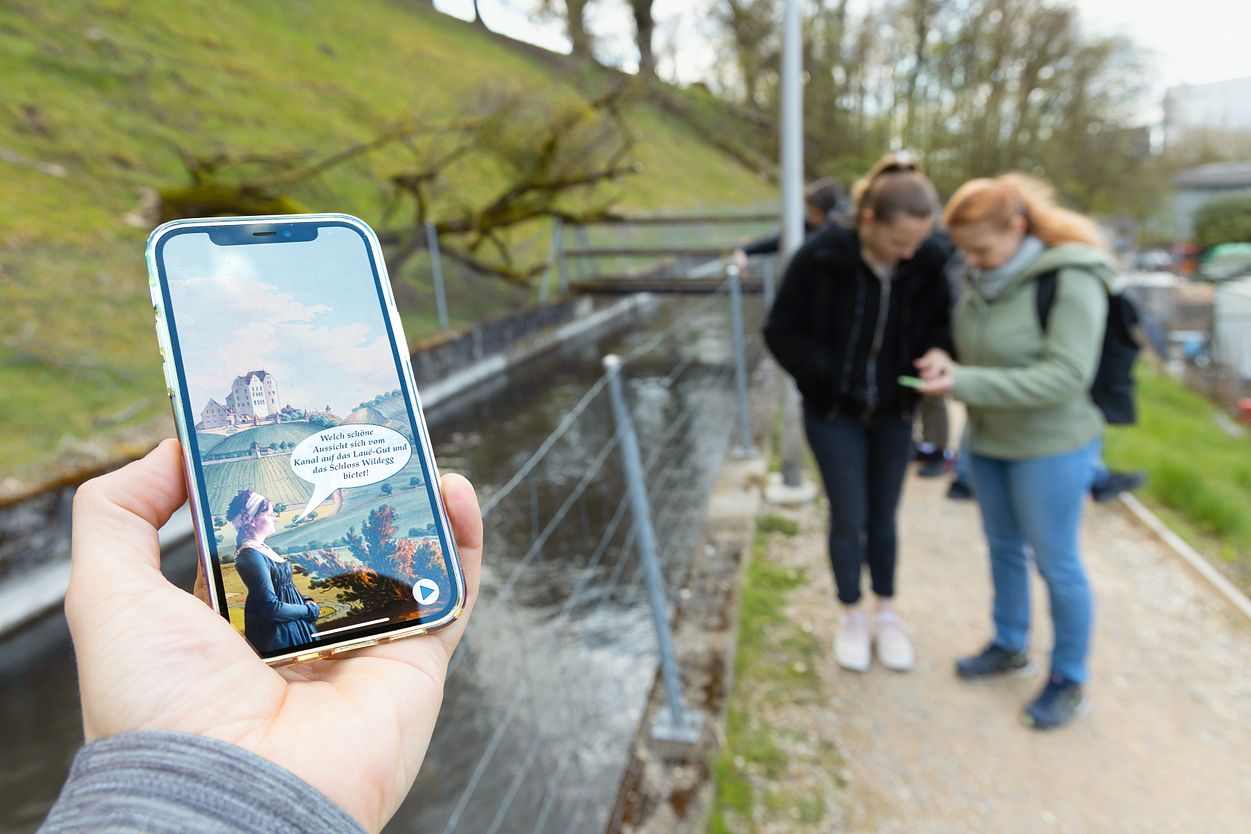 Nahaufnahme eines Mobiltelefons. Auf dem Screen ist die App "Industriekultur Aabach" sichtbar. Im Hintergrund sind mehrere Personen in Unschärfe zu sehen.