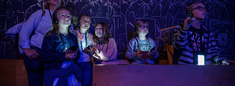 Eine Familie in einem dunklen Raum; die Displays der Audiogeräte um den Hals leuchten mystisch im Dunkeln. An der Rückwand Silhouetten von Legionären.
