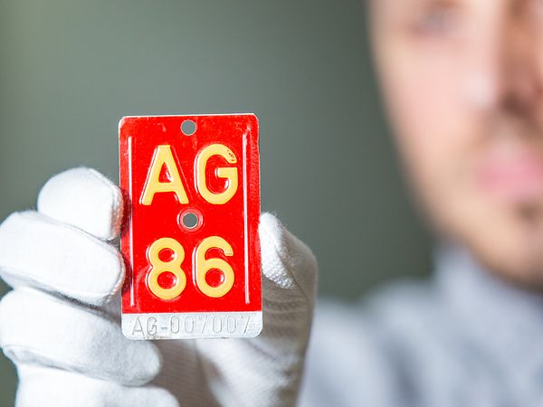 Rote Velonummer mit der Aufschrift "AG 86" in der Hand eines Mannes mit weissen Handschuhen. Im Hintergrund in Unschärfe der Kopf des Mannes (er hält die Velonummer in die Kamera).