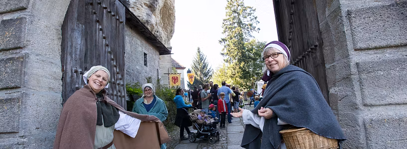 Zwei historisch gekleidete Freuen stehen vor dem Tor von Schloss Lenzburg und bitten Besucher mit einer einladenden Geste herein.