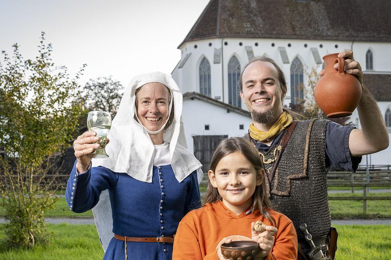 Eine mittelalterlich gekleidete Frau, ein römischer Legionär sowie ein modern gekleidetes Mädchen. Sie blicken alle freudig in die Kamera.