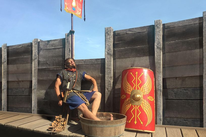 Ein Legionär sitzt auf einem Stuhl und blickt entspannt in die Sonne. Seine Füsse baden in einem Bottich mit Wasser.