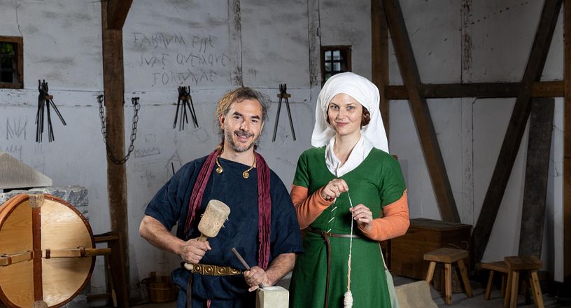 Ein römischer Steinmetz und eine mittelalterlich gekleidete Frau mit Wolle. Beide stehen in einem Werkraum und blicken direkt in die Kamera.