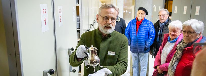 Ein Mann präsentiert im Sammlungszentrum Egliswil einen historischen Kelch. Neben und hinter dem Mann steht eine Besuchergruppe.