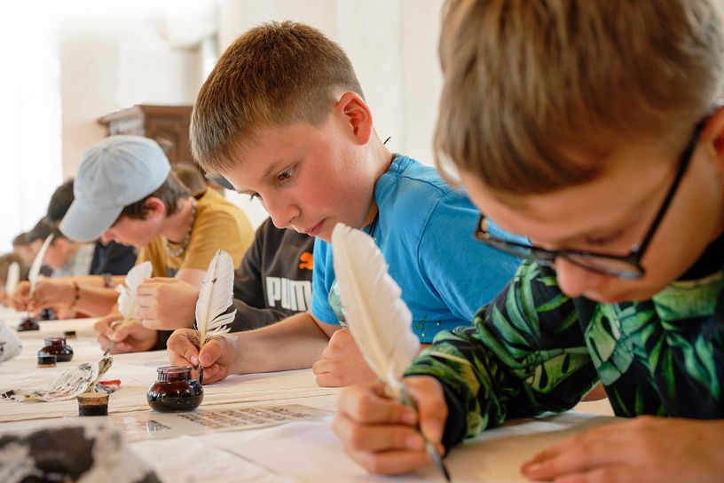 Mehrere Schüler und Schülerinnen schreiben mit einem Federkiel und Tintenfass auf Schloss Wildegg 