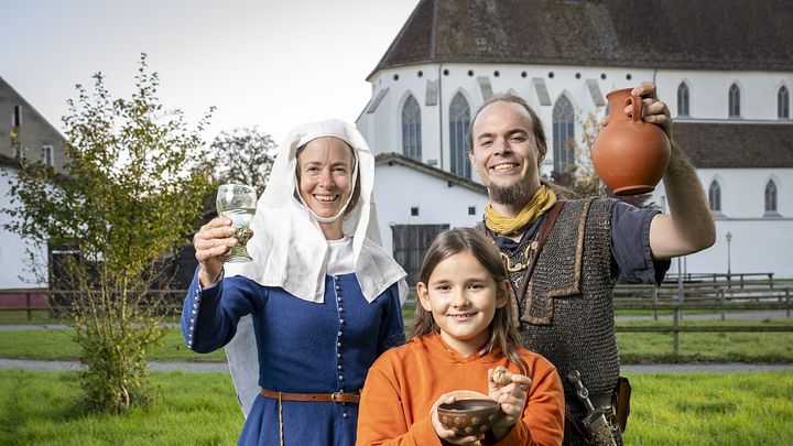 Eine mittelalterlich gekleidete Frau, ein römischer Legionär sowie ein modern gekleidetes Mädchen. Sie blicken alle freudig in die Kamera.