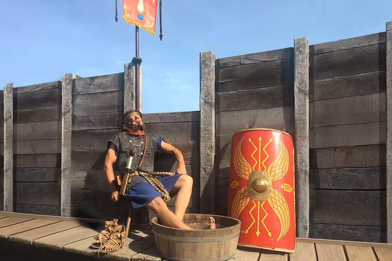 Ein Legionär sitzt auf einem Stuhl und blickt entspannt in die Sonne. Seine Füsse baden in einem Bottich mit Wasser.