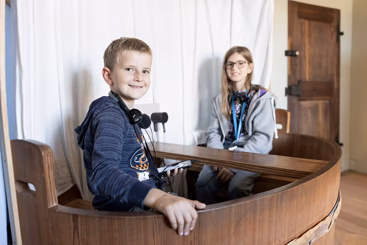 Ein Bub und ein Mädchen sitzen mit Kopfhörern in einem kleinen Schiff in der Ausstellung von Schloss Hallwyl. Der Bub blickt direkt in die Kamera.
