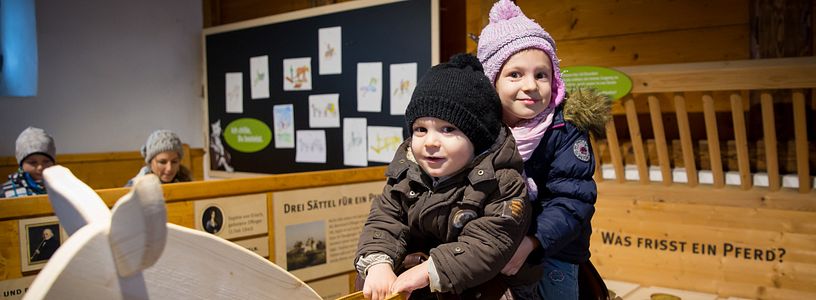 Mädchen und Junge reiten auf einem Holzpferd im Pferdestall auf Schloss Wildegg