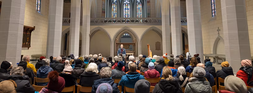 Innenraum Kloster Königsfeld mit vielen Gästen, die sitzen. Vorne spricht ein Mann an einem Podium.