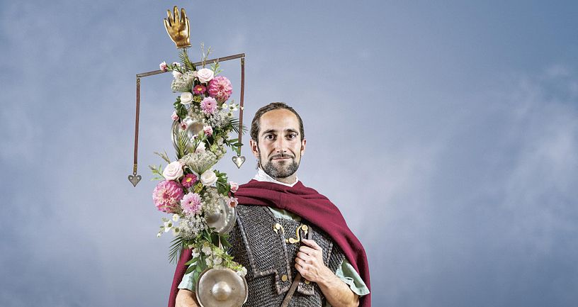 Ein römischer Legionär mit einer Standarte, die mit vielen Blumen geschmückt ist. Er blickt direkt in die Kamera. Der Hintergrund ist blau.