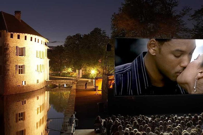 Nachtaufnahme von Schloss Hallwyl mit Aabach, rechts eine grosse Leinwand mit einer Filmszene (ein küssendes Paar).