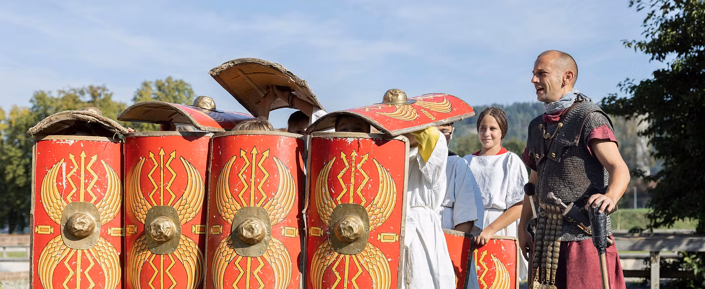 Gruppe von Kindern übt eine Formation mit Schilder, beaufsichtigt von einem Legionär im Legionärspfad Vindonissa