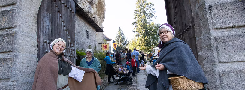 Zwei historisch gekleidete Freuen stehen vor dem Tor von Schloss Lenzburg und bitten Besucher mit einer einladenden Geste herein.