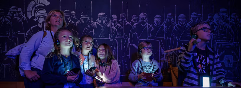 Eine Familie in einem dunklen Raum; die Displays der Audiogeräte um den Hals leuchten mystisch im Dunkeln. An der Rückwand Silhouetten von Legionären.
