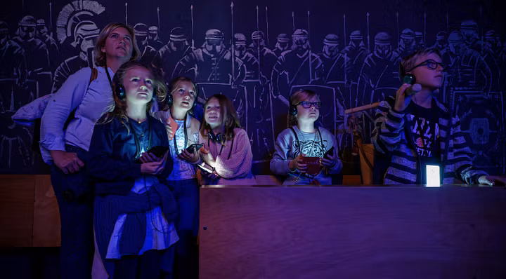 Eine Familie in einem dunklen Raum; die Displays der Audiogeräte um den Hals leuchten mystisch im Dunkeln. An der Rückwand Silhouetten von Legionären.