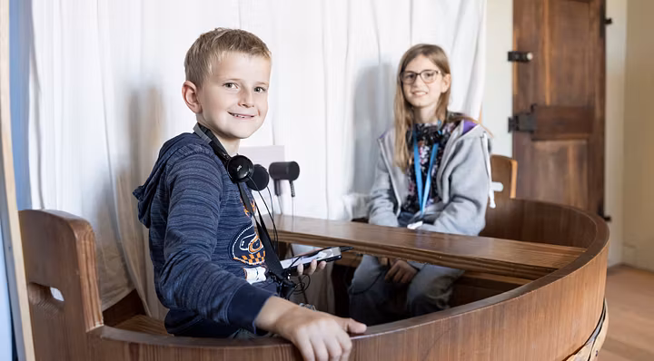 Ein Bub und ein Mädchen sitzen mit Kopfhörern in einem kleinen Schiff in der Ausstellung von Schloss Hallwyl. Der Bub blickt direkt in die Kamera.