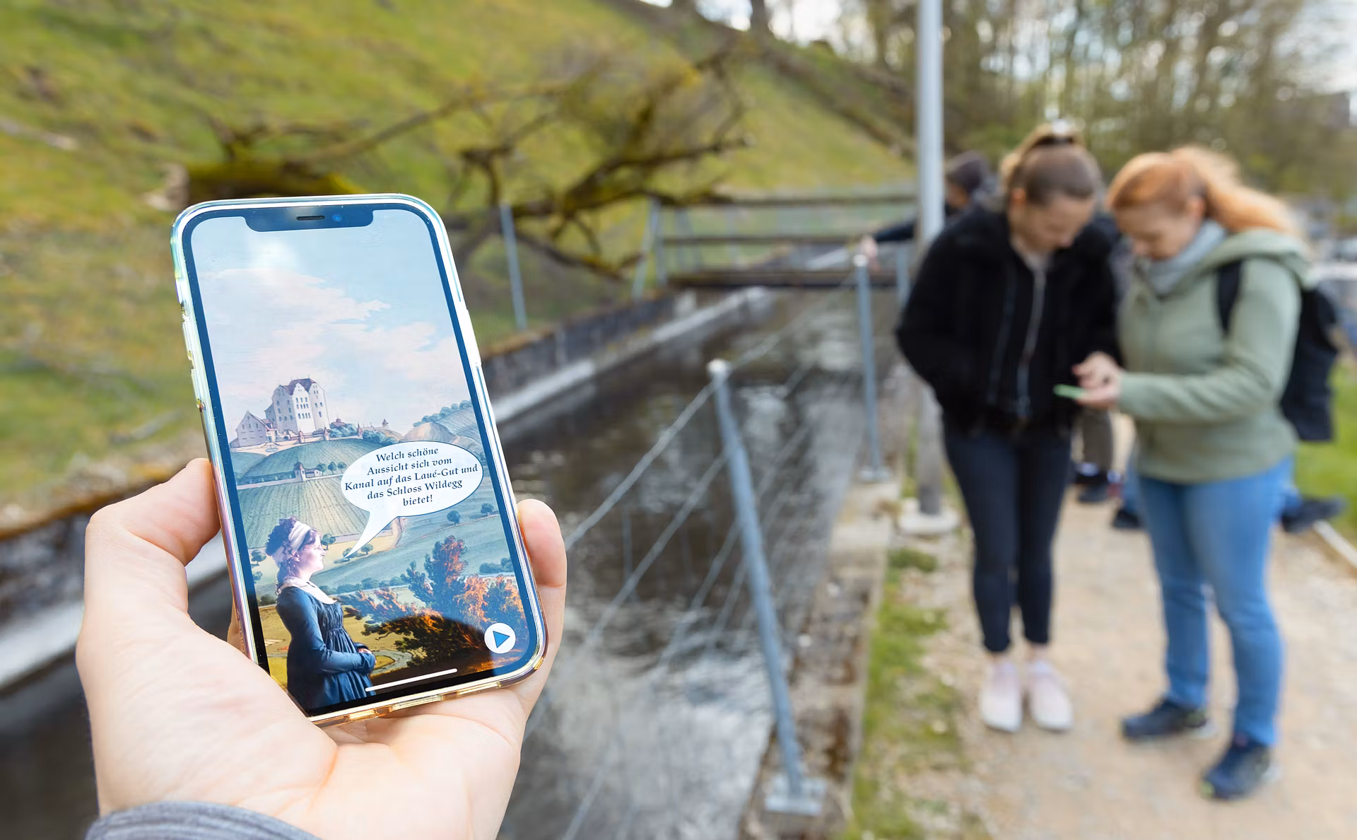 Nahaufnahme eines Mobiltelefons. Auf dem Screen ist die App "Industriekultur Aabach" sichtbar. Im Hintergrund sind mehrere Personen in Unschärfe zu sehen.