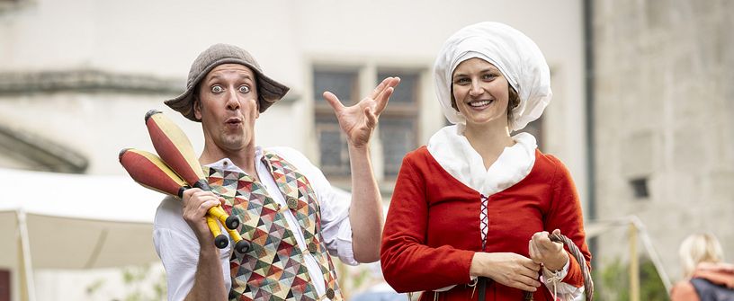 Eine mittelalterliche gekleidete Magd in rotem Kleid, neben ihr steht ein Gaukler mit Keulen. Er macht Faxen.