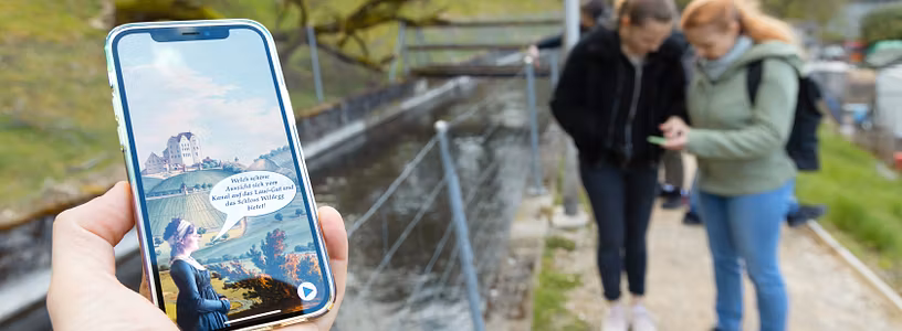 Nahaufnahme eines Mobiltelefons. Auf dem Screen ist die App "Industriekultur Aabach" sichtbar. Im Hintergrund sind mehrere Personen in Unschärfe zu sehen.