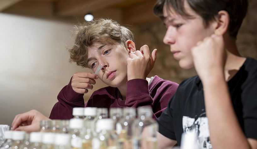 Ein Schüler riecht an einem Duftstäbchen, vor ihm steht eine Duftorgel mit diversen verschiedenen Dürfen in kleinen Flaschen.