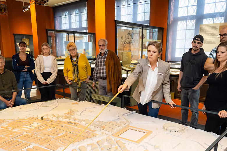 Eine Museumsführerin sowie mehrere Personen bei einer Führung im Vindonissa Museum. Die Gruppe steht um ein grosses Modell des Legionionslagers Vindonissa. Die Führerin zeigt mit einem Holzstab auf das Modell.