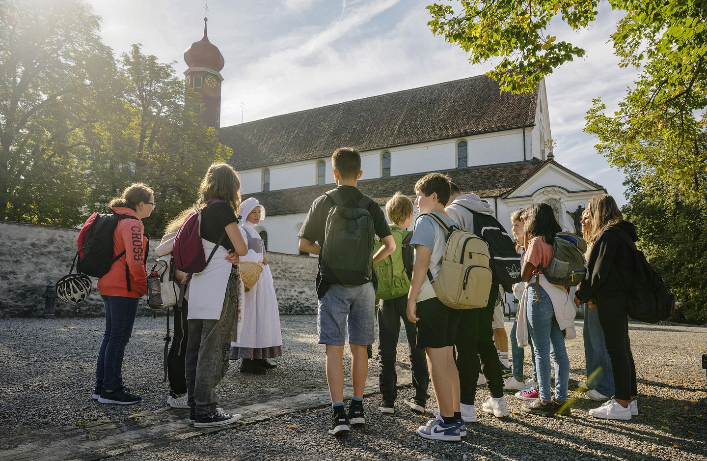 Eine Gruppe Schülerinnen und Schüler steht auf dem Vorplatz der Klosterhalbinsel Wettingen. Sie hören einer Klostermagd im Kostüm zu.