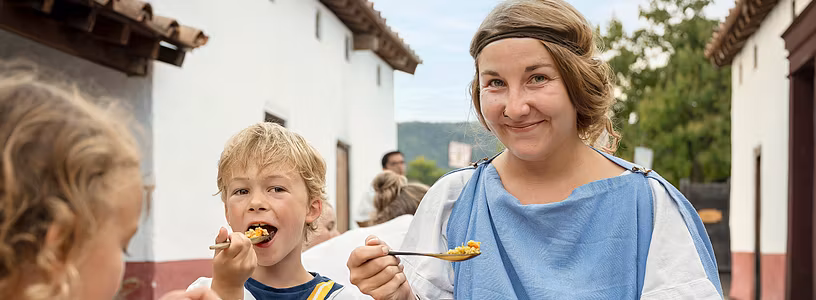 Eine römisch gekleidete Frau und ein römisch gekleideter Bub sitzen an einem Tisch und essen gemeinsam. Sie blicken direkt in die Kamera.