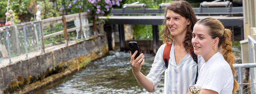 Zwei Frauen stehen an einem Bach, sie halten Handy in den Händen.
