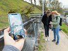 Nahaufnahme eines Mobiltelefons. Auf dem Screen ist die App "Industriekultur Aabach" sichtbar. Im Hintergrund sind mehrere Personen in Unschärfe zu sehen.