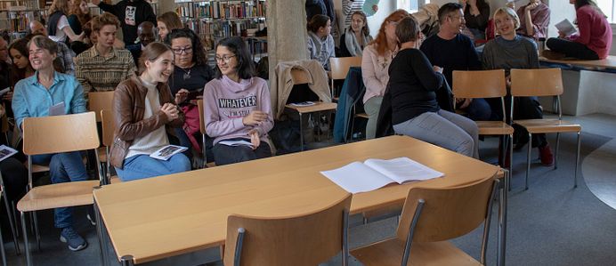 Ganz vorne ist ein Tisch mit zwei Stühlen gegen ein Publikum gerichtet, das aus mehreren Jugendlichen und Erwachsenen besteht. Gewisse Personen stehen im Hintergrund noch. Dahinter sind Bücherregale zu sehen, es handelt sich um eine Bibliothek. In der Mitte des Raumes gibt es vier Steinsäulen, die sich von vorne bis hinten ziehen.