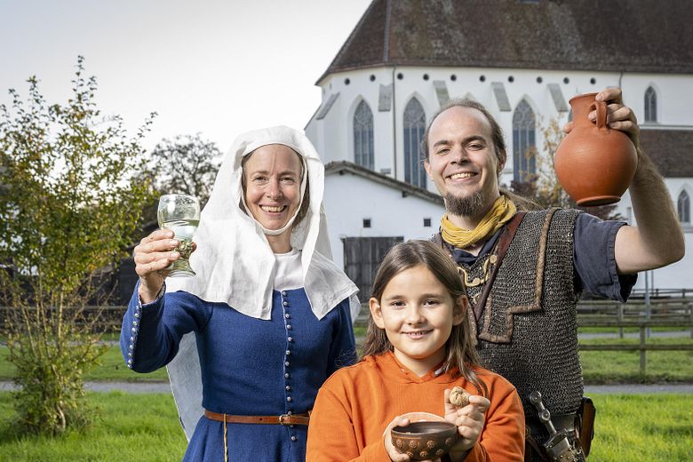 Eine mittelalterlich gekleidete Frau, ein römischer Legionär sowie ein modern gekleidetes Mädchen. Sie blicken alle freudig in die Kamera.