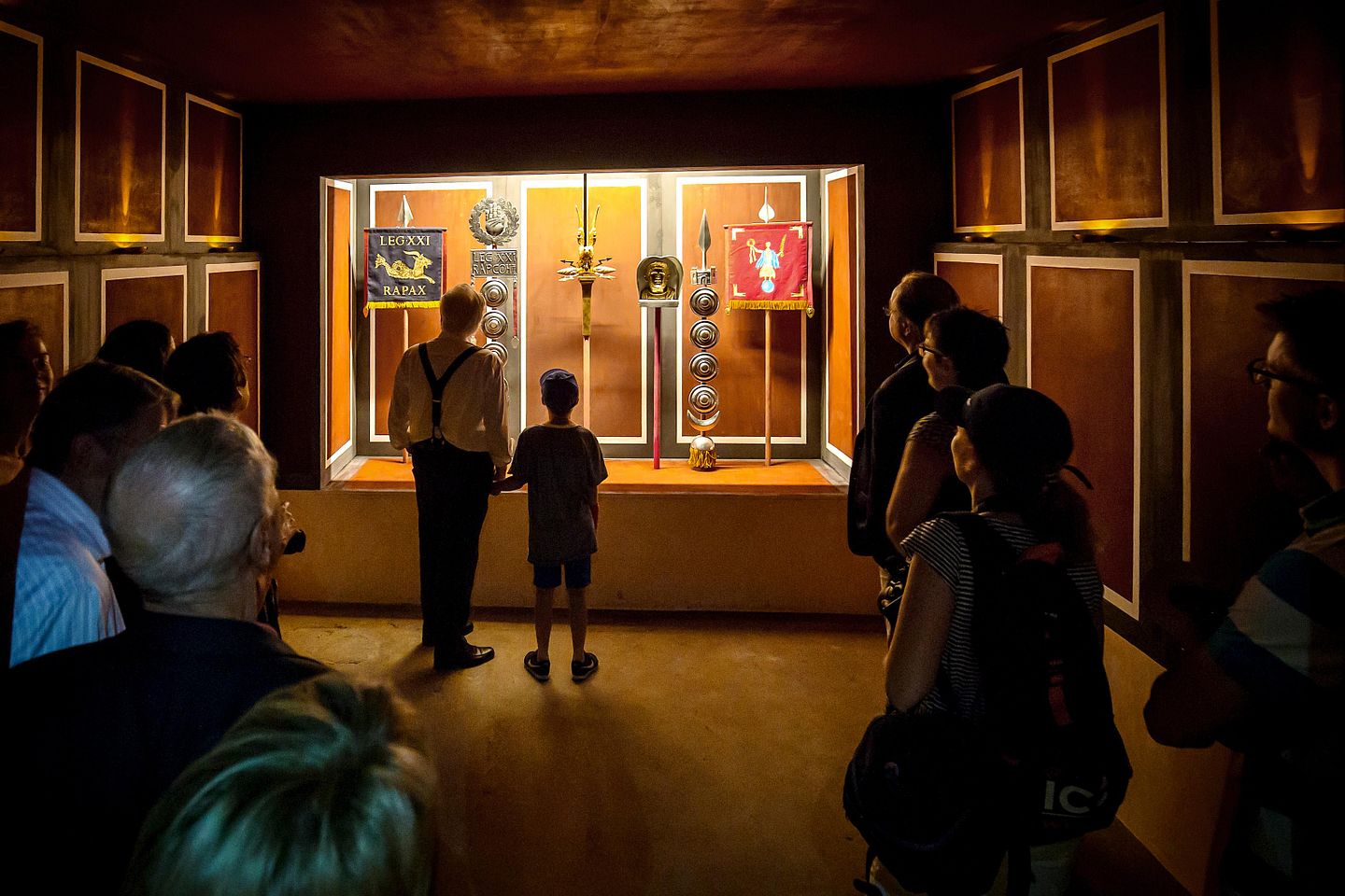 Ein stimmungsvoll erleuchteter Raum mit römischen Objekten in einer Vitrine, davor stehen mehrere Personen.