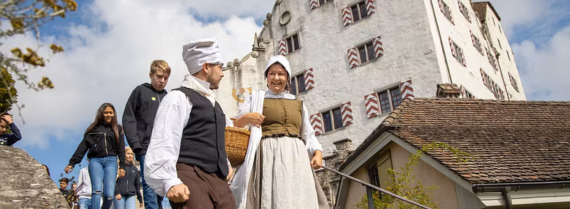 Eine Magd und ein Knecht vor Schloss Wildegg, dahinter eine Schulklasse.