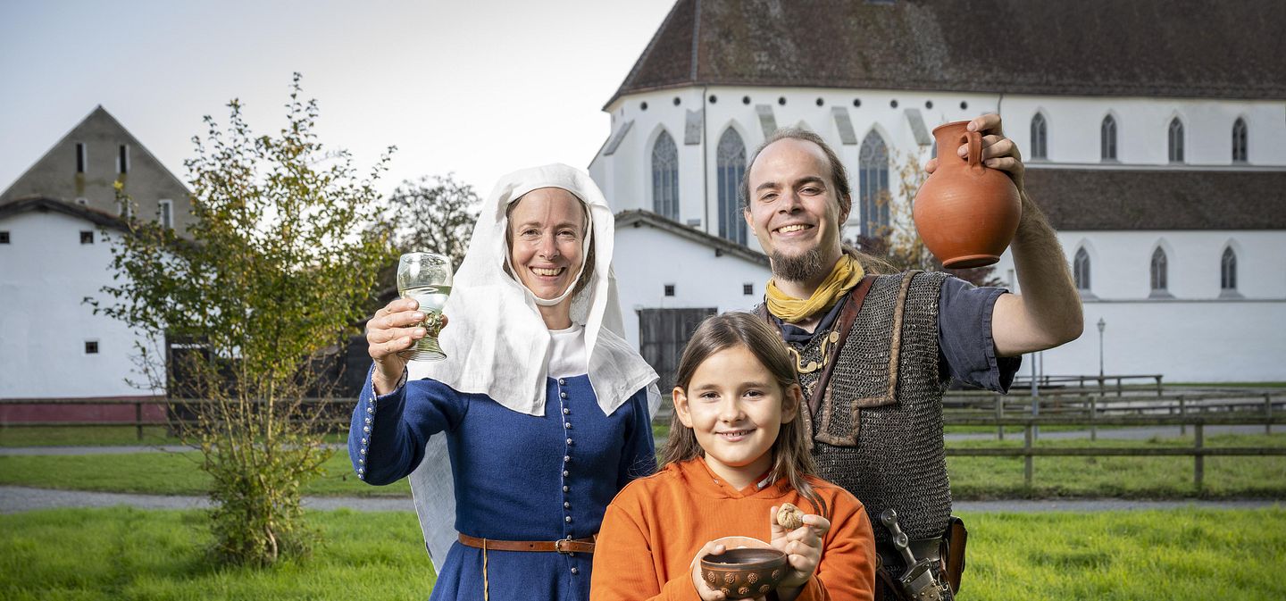 Eine mittelalterlich gekleidete Frau, ein römischer Legionär sowie ein modern gekleidetes Mädchen. Sie blicken alle freudig in die Kamera.