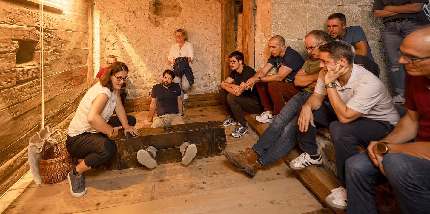 Eine Gruppe Männer und Frauen sitzen mit einer Museumsführerin im Gefängnis von Schloss Lenzburg. Eine Person liegt am Boden, die Füsse sind in einem Holz fixiert