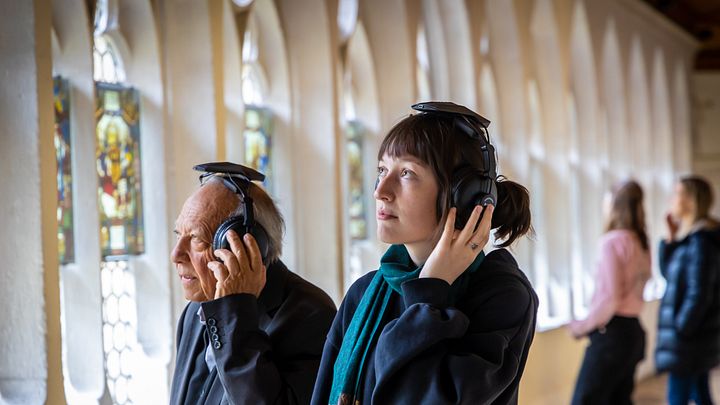 Ein Mann und eine Frau stehen im Kreuzgang der Klosterhalbinsel Wettingen. Beide tragen grosse Kopfhörer. Im Hintergrund weitere Personen.