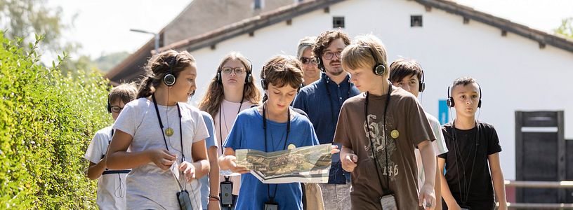 Eine Schulklasse unterwegs auf einem Kiesweg; im Hintergrund die rekonstruierte Unterkunft von Legionären.
