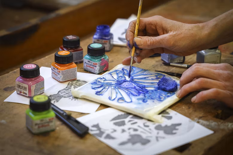Nahaufnahme einer Hand, die mit Pinsel und Farben eine weisse Kacheln bemalt.