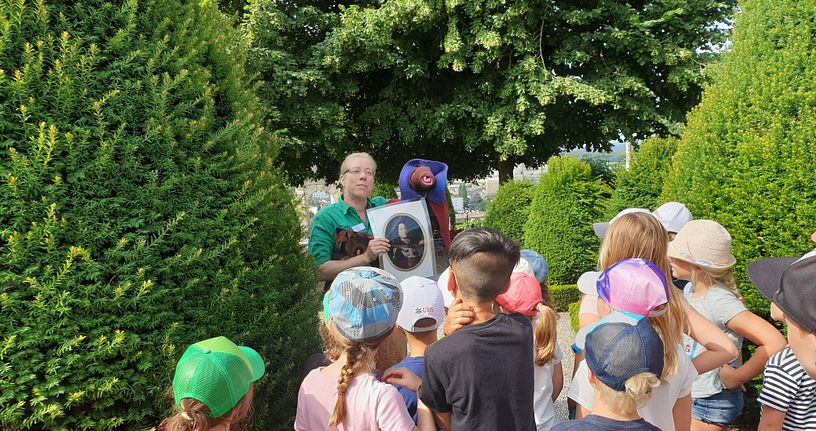 Eine Frau steht mit einer Handpuppe im Garten von Schloss Wildegg, vor ihr eine Gruppe Schülerinnen und Schüler (Führung)