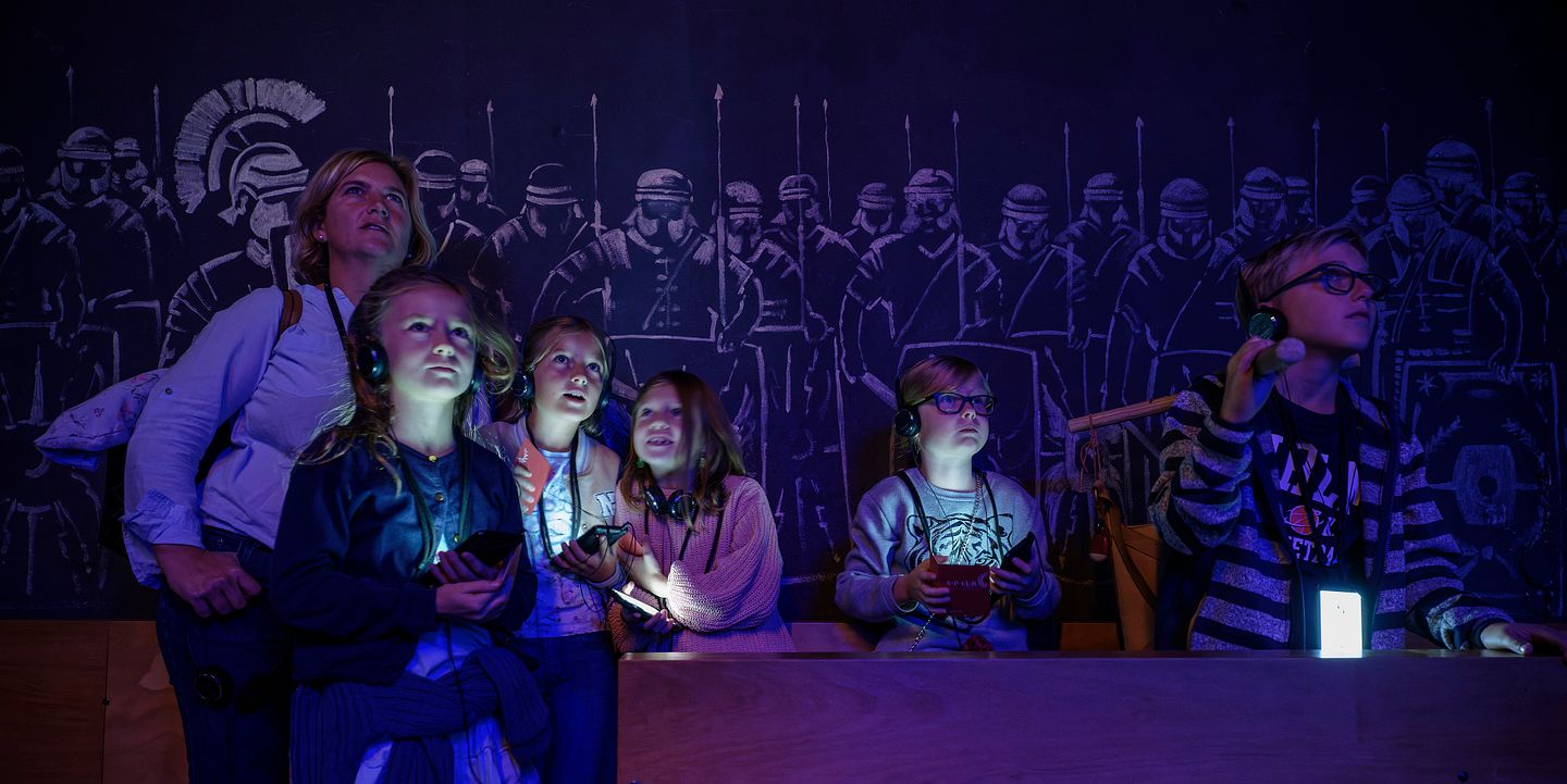 Eine Familie in einem dunklen Raum; die Displays der Audiogeräte um den Hals leuchten mystisch im Dunkeln. An der Rückwand Silhouetten von Legionären.