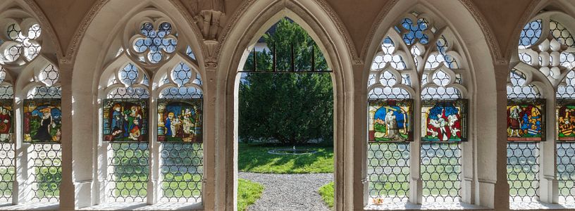 Frontaler Blick auf Glasfenster im Kreuzgang des Kloster Wettingen. In der Mitte führt ein grosser Durchgang in den dahinterliegenden Kreuzganggarten.