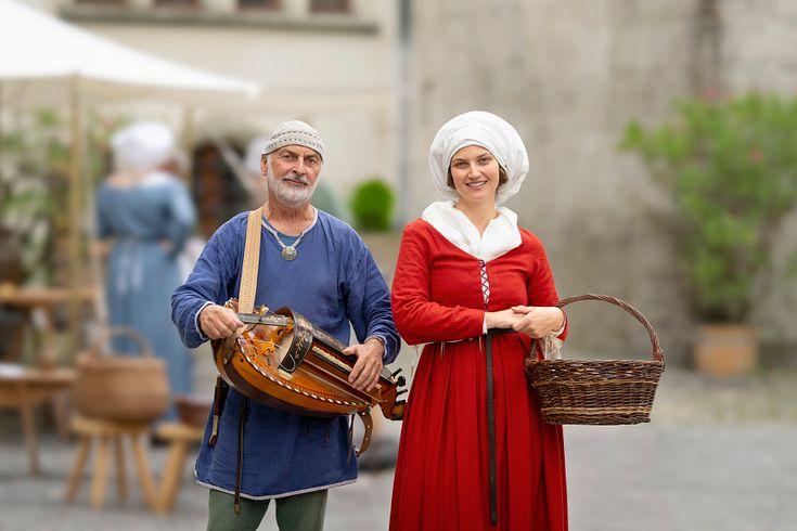 Eine mittelalterlich gekleidete Hofdame mit einem Musiker (mit Drehleier in der Hand). Beide blicken direkt in die Kamera.