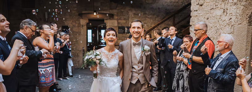 Ein Hochzeitspaar auf Schloss Habsburg: Ein Mann und eine Frau schreitet durch spalierstehende Gäste. Seifenblasen fliegen in der Luft.