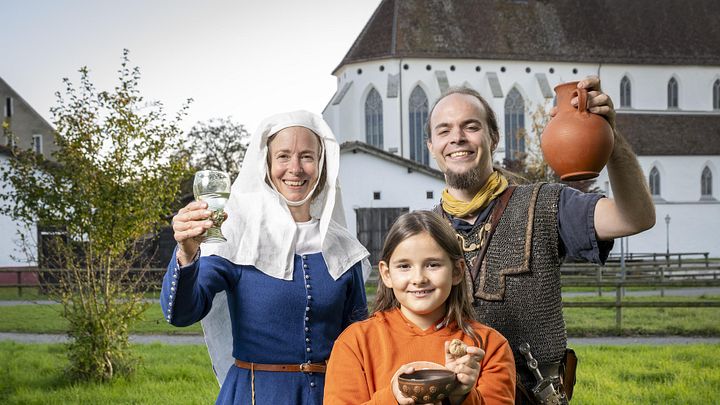 Eine mittelalterlich gekleidete Frau, ein römischer Legionär sowie ein modern gekleidetes Mädchen. Sie blicken alle freudig in die Kamera.