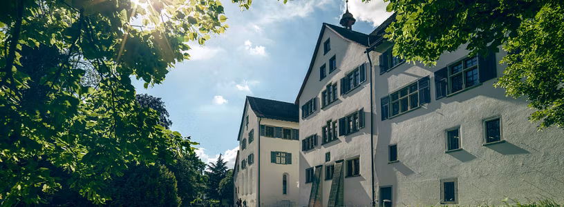 Aussenaufnahme Kloster Wettingen mit blauen Himmel, Sonne im Gegenlicht und einer grünen Wiese im Vordergrund.
