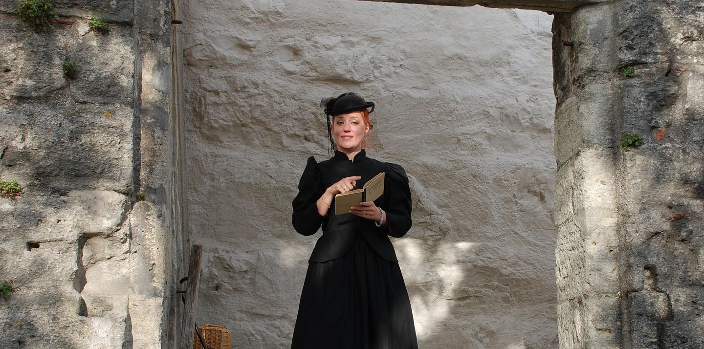 Eine als Gouvernante verkleidete Frau mit Buch auf Schloss Wildegg (blickt direkt in die Kamera)