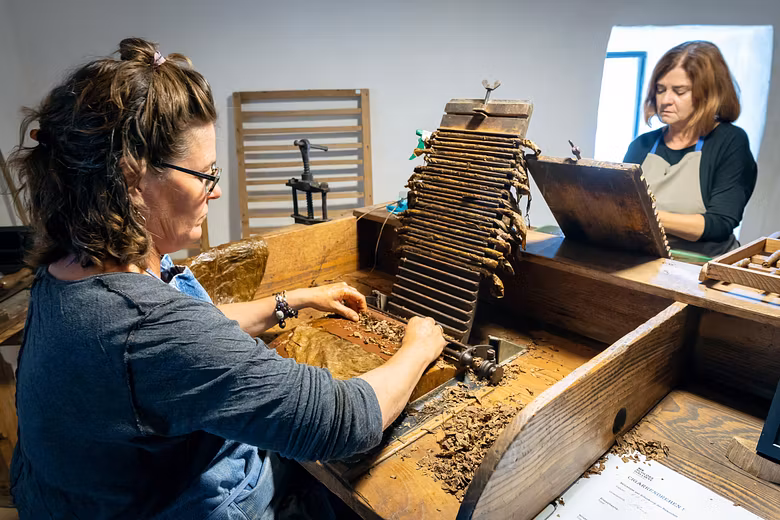 Zwei Frauen sitzen an einem Holztisch und drehen aus Tabak Zigarren.