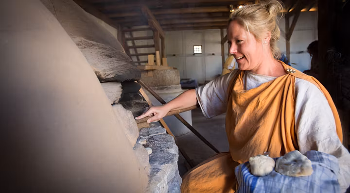 Eine römisch gekleidete Frau holt Brot aus einem römischen Steinofen im Legionärspfad Vindonissa.
